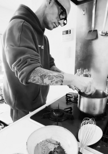 Brendan, founder of Azabu Foods, is seen in a black-and-white photo experimenting in the kitchen. Wearing a hoodie and glasses, he carefully stirs ingredients in a saucepan on a gas stove, fine-tuning the recipe for Umami Everything. A bowl with spices and sesame seeds sits on the counter, along with a whisk, capturing the behind-the-scenes process of product development.